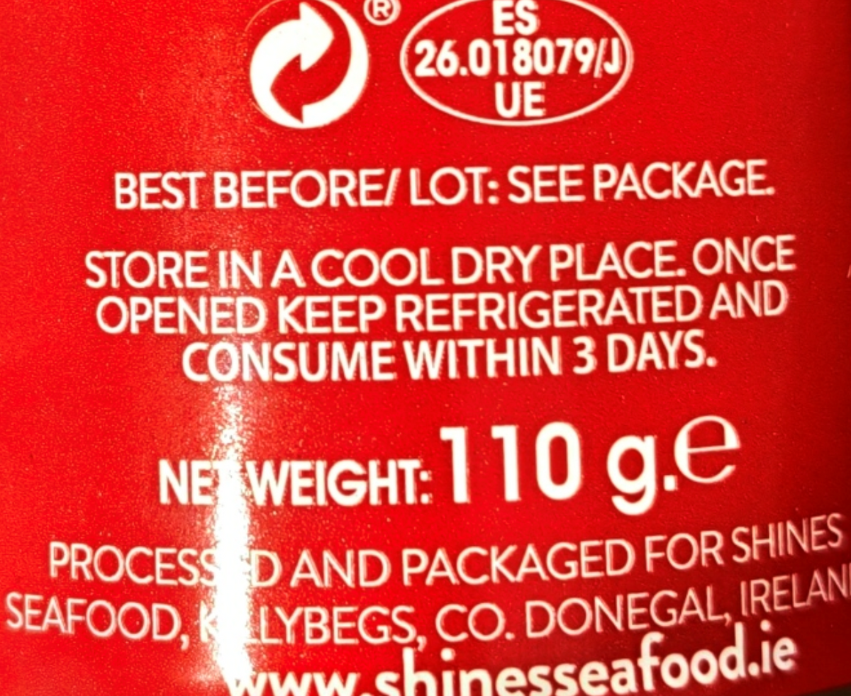 Close-up of a red food product package showing white printed text with storage instructions, a recycling symbol, ES/UE codes, net weight 110 g, and the website shinesseafood.ie.