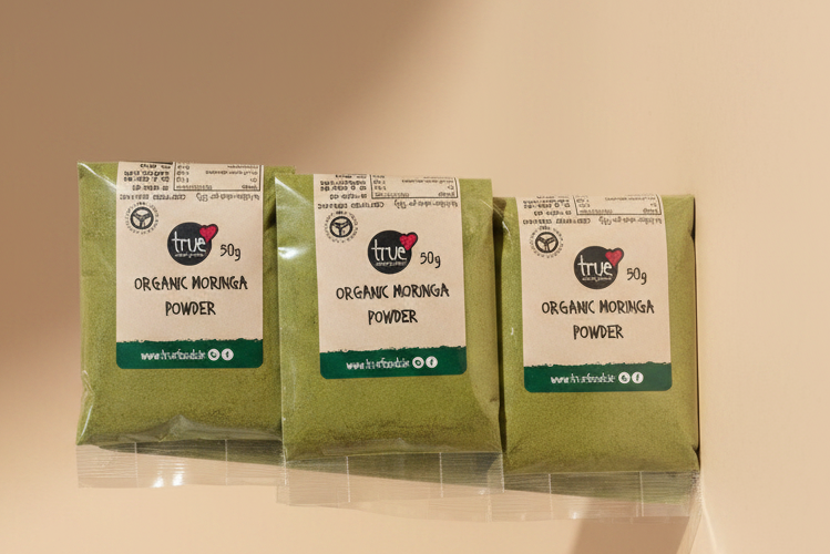 Three bags of True Organic Morinda Powder stacked vertically inside a clear plastic outer bag, showing the green powder and white labels.