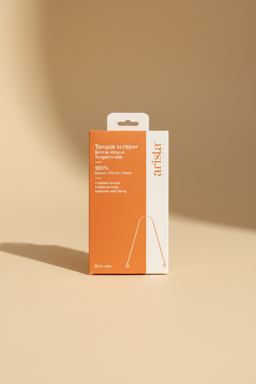 A hand holding a box of Arista Copper Tongue Scraper against a blurred indoor background. The packaging is orange and white with text describing it as a 100% copper oral care tool.