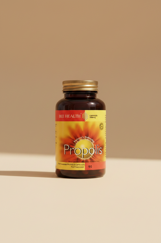A bottle of Bee Health Propolis food supplement capsules, held in a hand. The bottle is dark amber brown with a gold cap, and the label features a yellow and orange bee-flower design with the brand name 'BEE HEALTH' and product name 'Propolis' in white text.