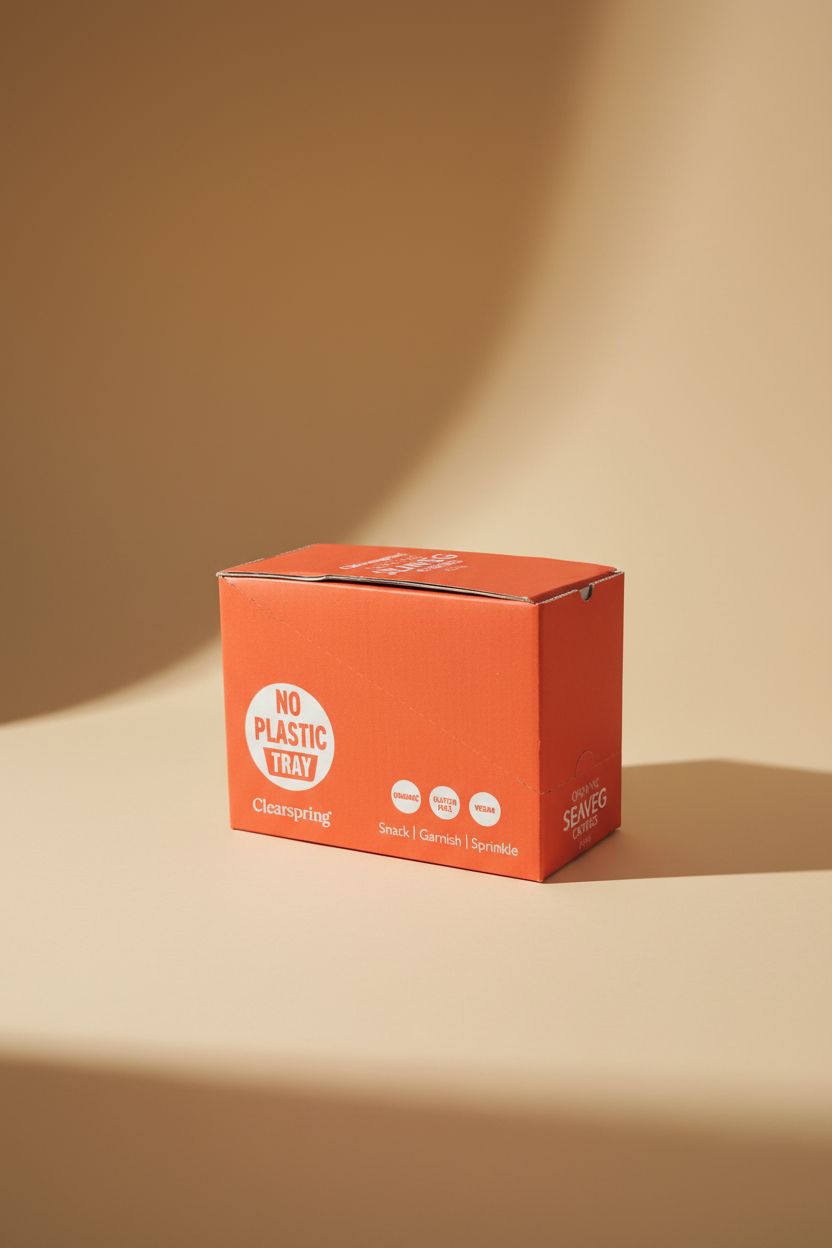 A red cardboard box for Clearspring snack, garnish, and sprinkle, featuring a white circular logo that reads 'NO PLASTIC TRAY' and icons for organic, gluten free, and vegan. The box is photographed on a cardboard surface in a warehouse or storage setting.