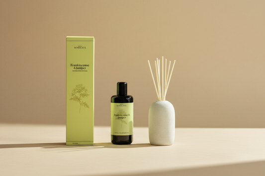 A reed diffuser set from The Herbalists, featuring a black bottle of Frankincense & Juniper oil, a tall light green box, and a white ceramic reed diffuser vase with wooden reeds, styled with moss, pinecones, and a rustic brown background.