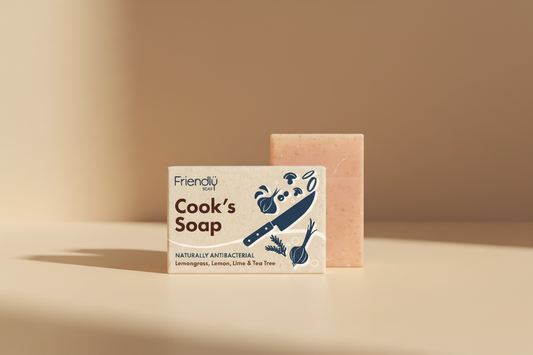 A bar of soap labeled 'Friendlü Cook's Soap' shown with its printed packaging card and the soap bar behind it on a white background. The soap is peachy pink with small speckles, and the card features dark blue and brown text with illustrations of a knife, garlic, onion, and herbs.