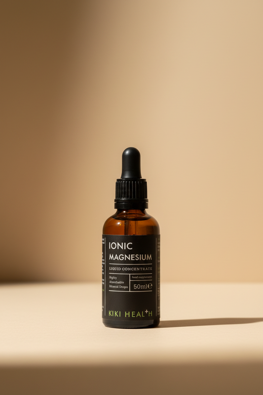 A hand holding a 50ml amber glass dropper bottle of Kiki Health Ionic Magnesium Liquid Concentrate against a blurred indoor background.