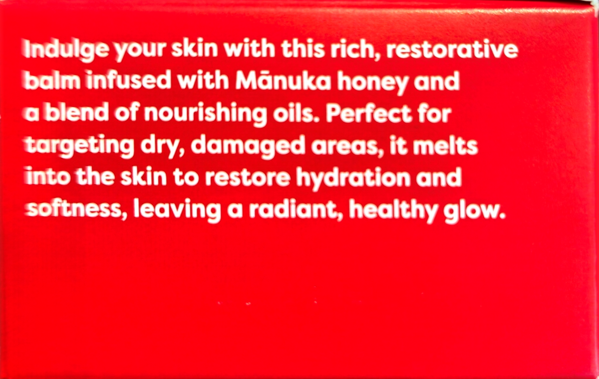 A close-up of a bright red box or package with white text describing a skin balm infused with Mánuka honey and nourishing oils.