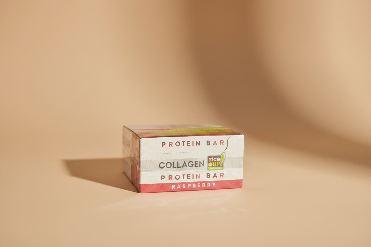 A hand holding a shrink-wrapped box of Collagen Protein Bar in Raspberry flavor. The packaging features a red and white design with a yellow and green Ice Dri logo, wrapped in clear plastic.