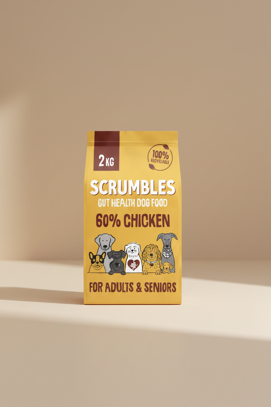 A 2 kg bag of Scrumbles Gut Health Dog Food, 60% Chicken, for adults and seniors. The packaging is a warm golden yellow with brown and white text, featuring cartoon illustrations of various dog breeds at the bottom.