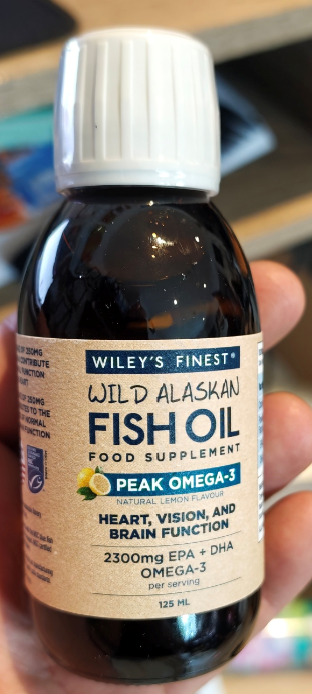 A hand holding a bottle of Wiley's Finest Wild Alaskan Fish Oil Food Supplement. The bottle is dark amber glass with a white ribbed cap and a beige label featuring blue and green text.