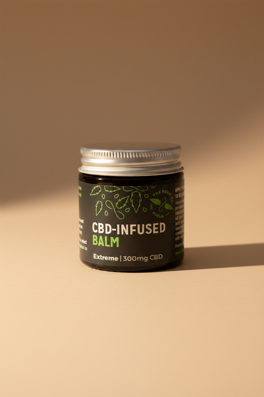 A jar of CBD-infused balm with a silver metal lid and a black label featuring green leaf illustrations and white and green text. The label reads 'CBD-INFUSED BALM' and 'Extreme | 300mg CBD'.